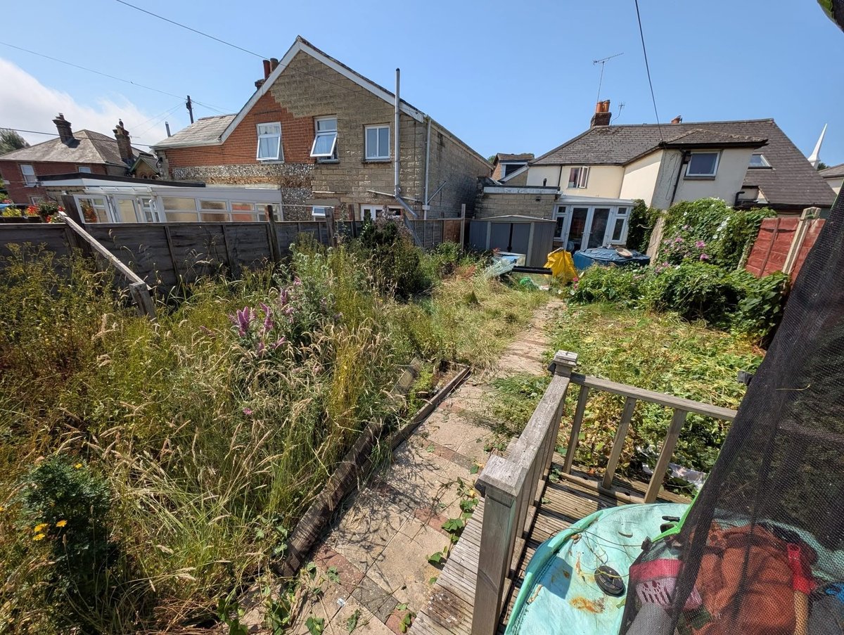 Overgrown residential garden with stone cottages, wooden fence, and childrens toys visible in a rural village setting