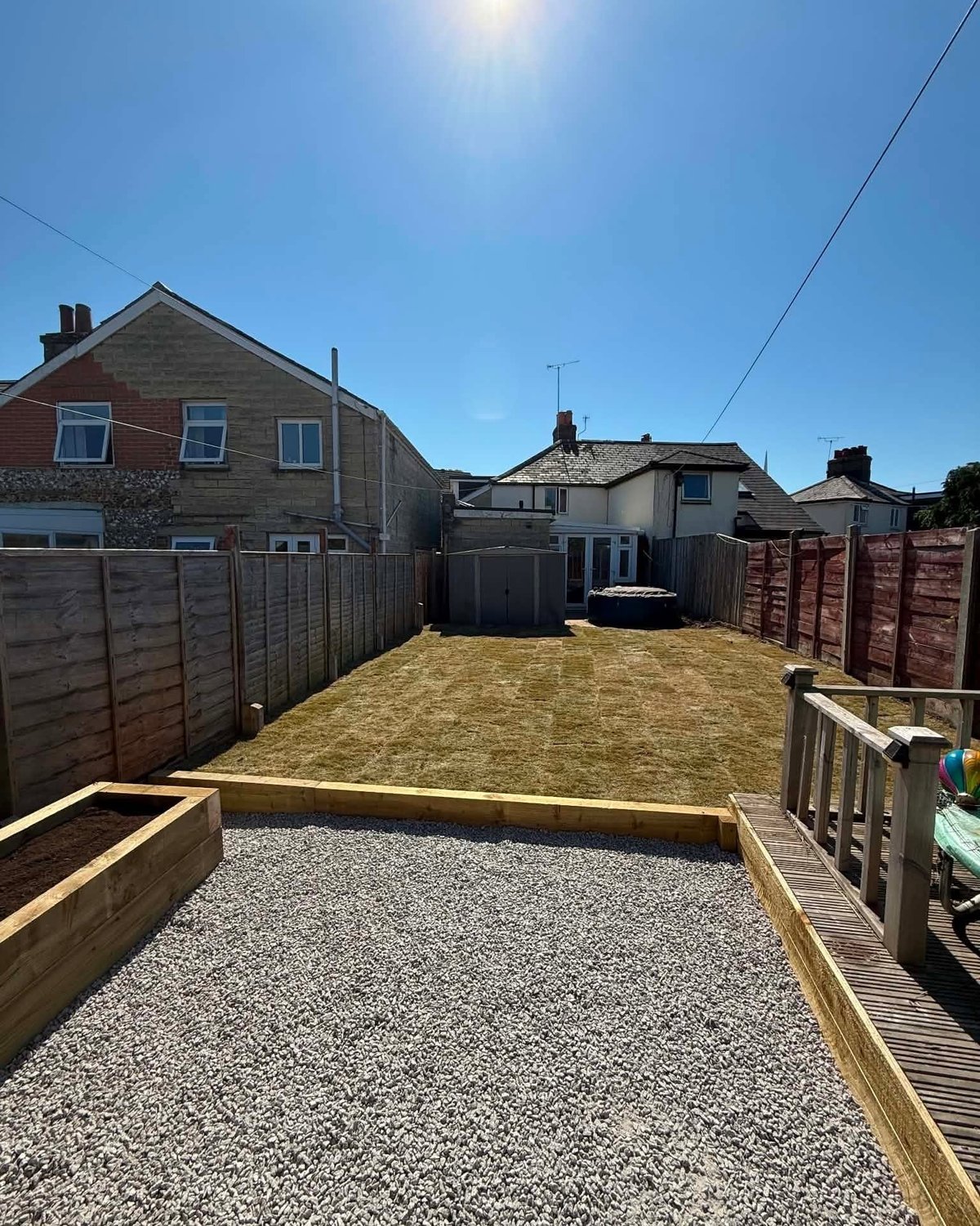 Residential backyard with gravel patio, wooden fences, brick houses, and clear blue sky in background