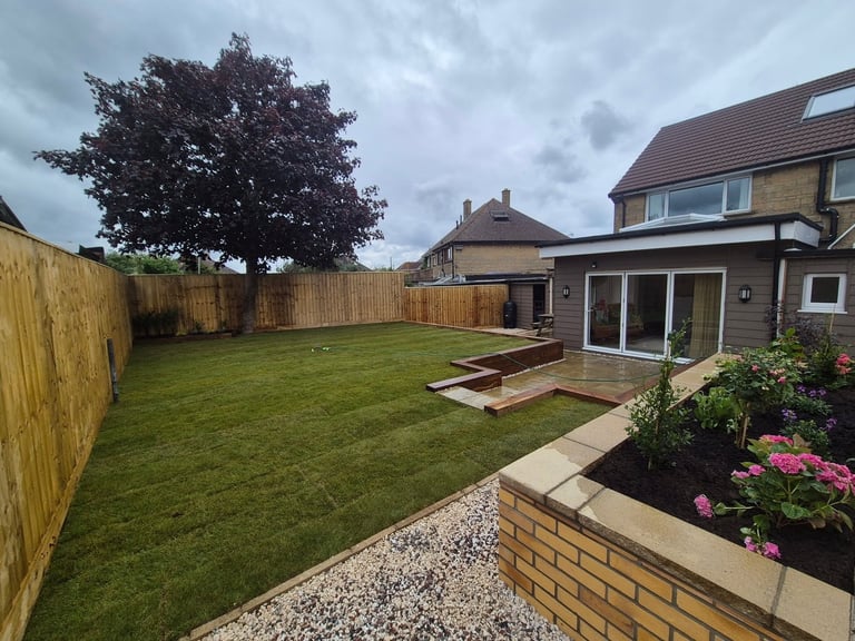 Spacious residential backyard with green lawn, brick patio, wooden deck, flowering plants, and brown wooden fence under cloudy sky